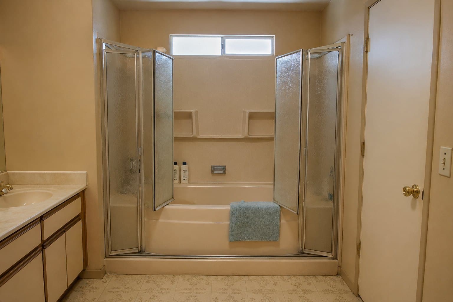 An outdated 1990s Arizona primary bathroom — almond-beige built-in fiberglass tub, dated almond fiberglass surround panels (not real tile), cloudy white vinyl bi-fold doors, warm tan walls, and a beige laminate vanity. Clean but slippery and overdue for a refresh.